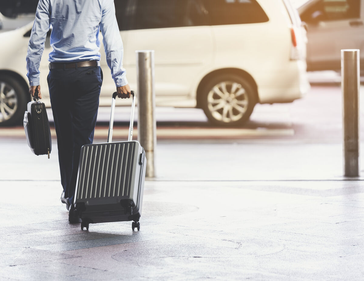 A traveller wheeling a suitcase past a waiting vehicle at a terminal kerb