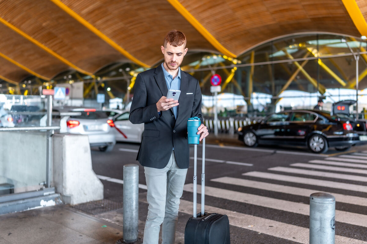 Traveller with a suitcase checking a phone at an airport kerbside pickup